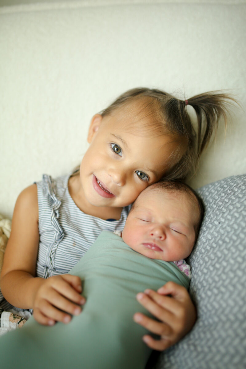 A proud big sister snuggling her newborn brother in a sage swaddle