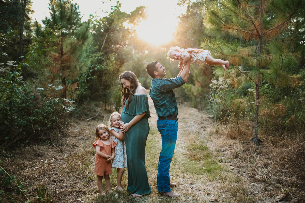 An expecting family of five laughing and playing together in a sunlit pine clearing