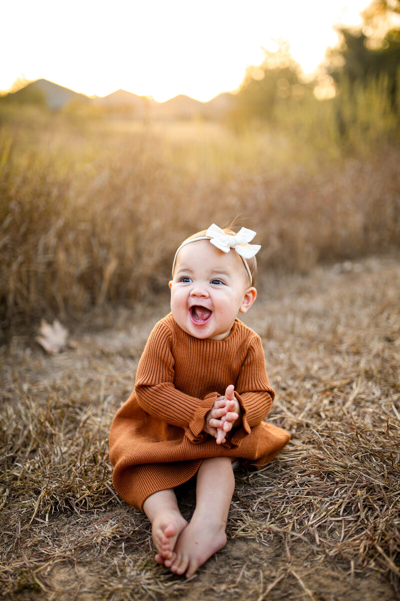 A laughing baby girl in a rust knit dress sitting in a golden field at sunset