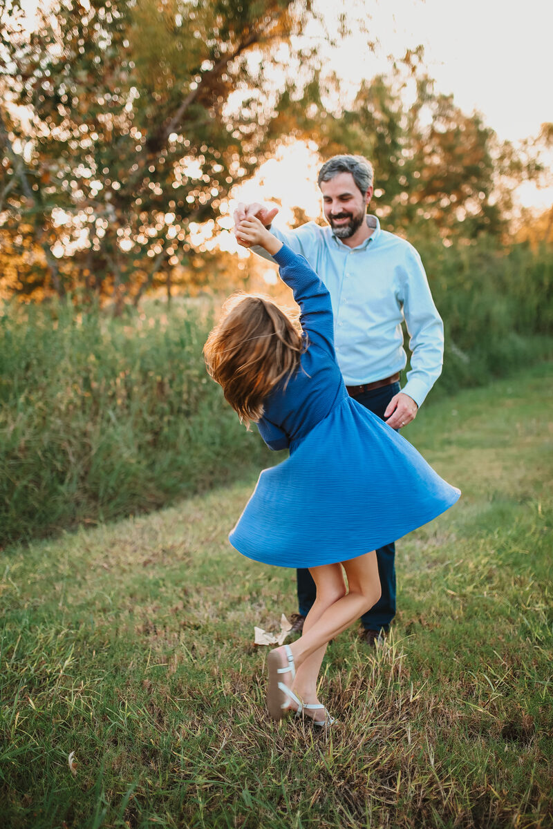 A father twirling his daughter in a blue dress in a sunlit field