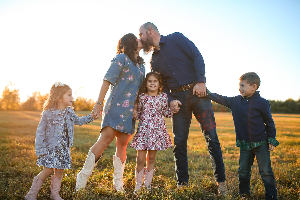 Parents sharing a kiss while their three children hold hands in a sunset field