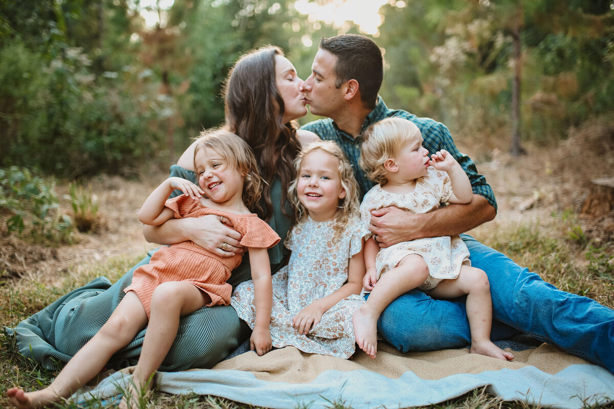 A family cuddled on a blanket, parents sharing a kiss as their three girls giggle
