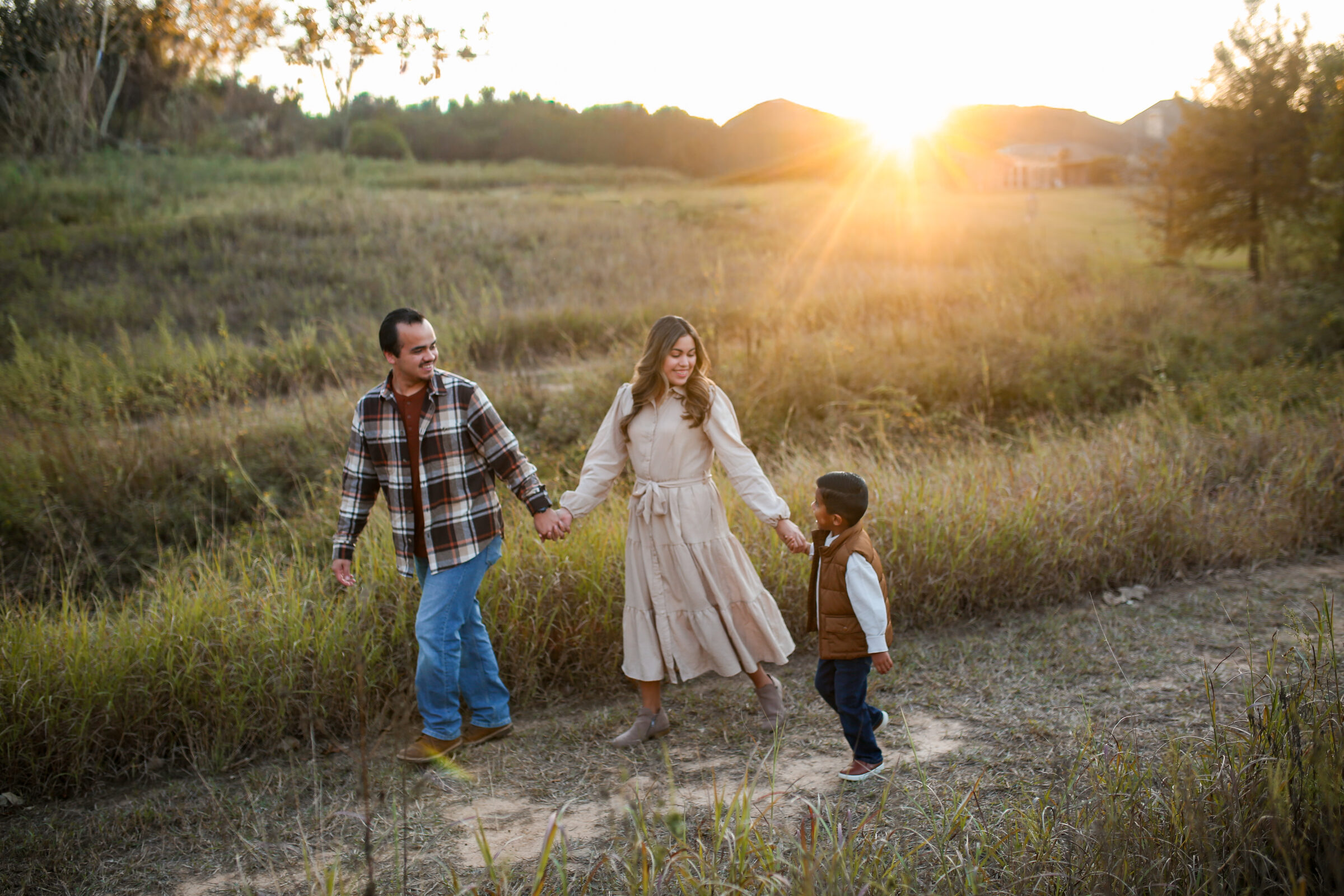 A mother, father, and young son walking hand in hand through a golden field at sunset
