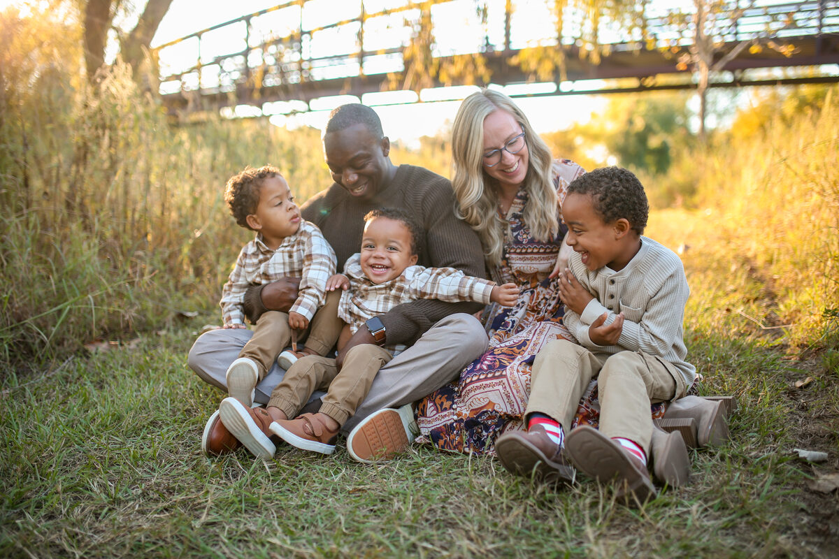 A family of five snuggled together in golden grass beneath a footbridge