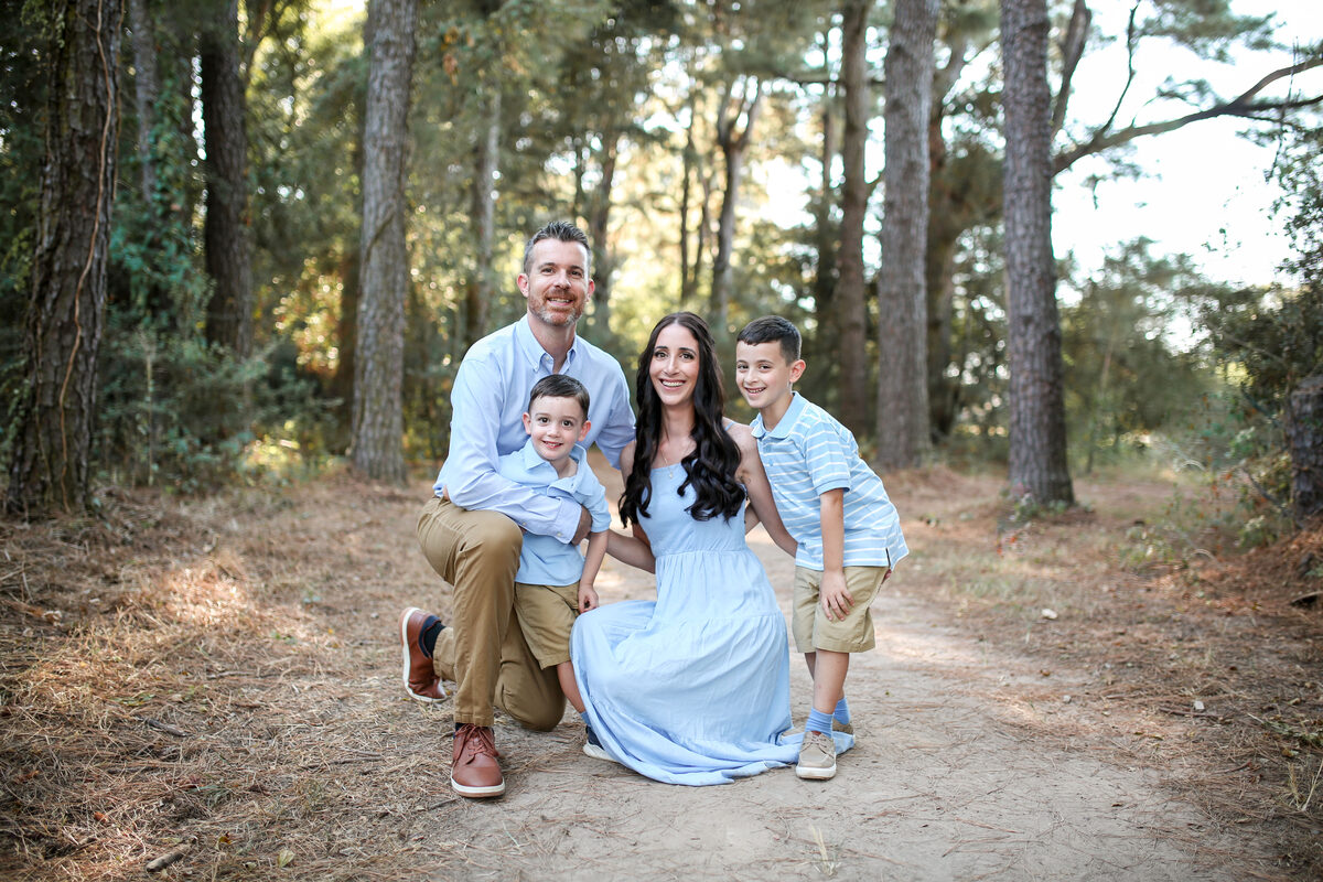 A family of four in soft blues kneeling together on a pine path