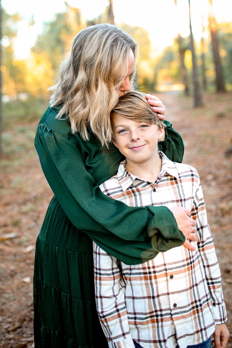 A mother kissing her tween son's head on a golden pine trail