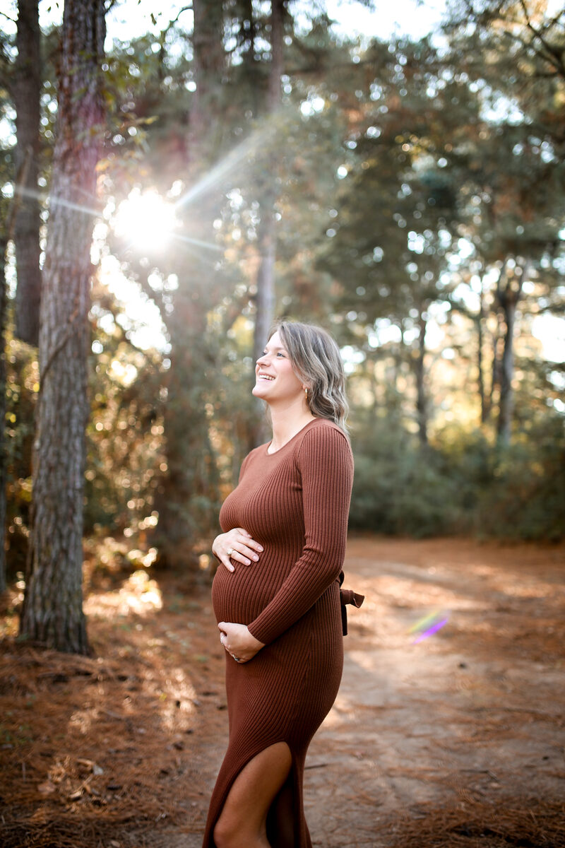 An expecting mother in a rust dress smiling in the sunlit pines