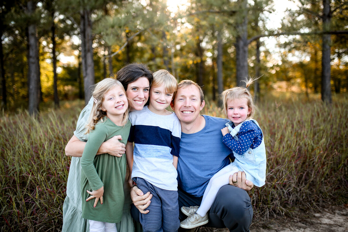 A family of five smiling together in tall grass at the edge of a pine forest