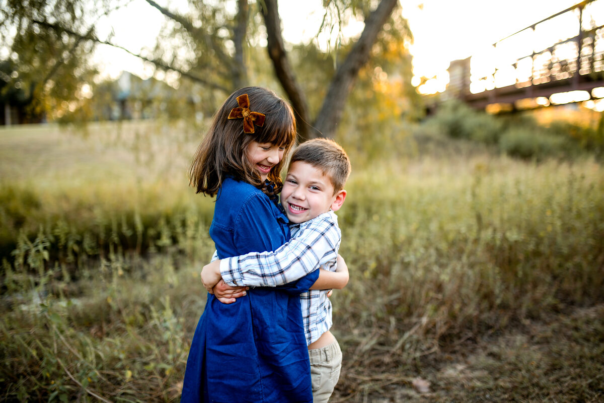 A brother and sister hugging in tall grass near a wooden footbridge