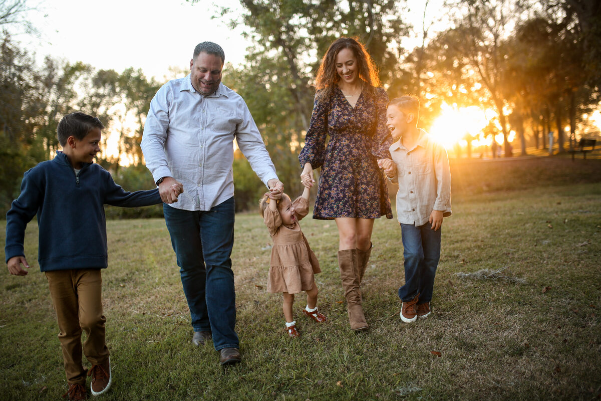A family of five walking hand in hand through a sunset park, swinging their toddler