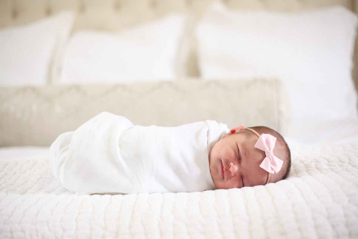A newborn girl with a pink bow swaddled on a white bed
