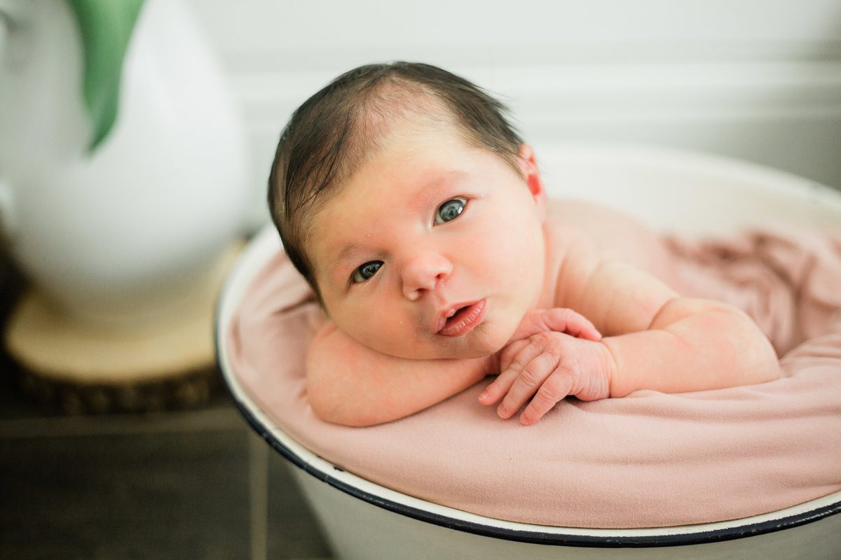 A wide-eyed newborn resting her chin on the edge of a vintage tub
