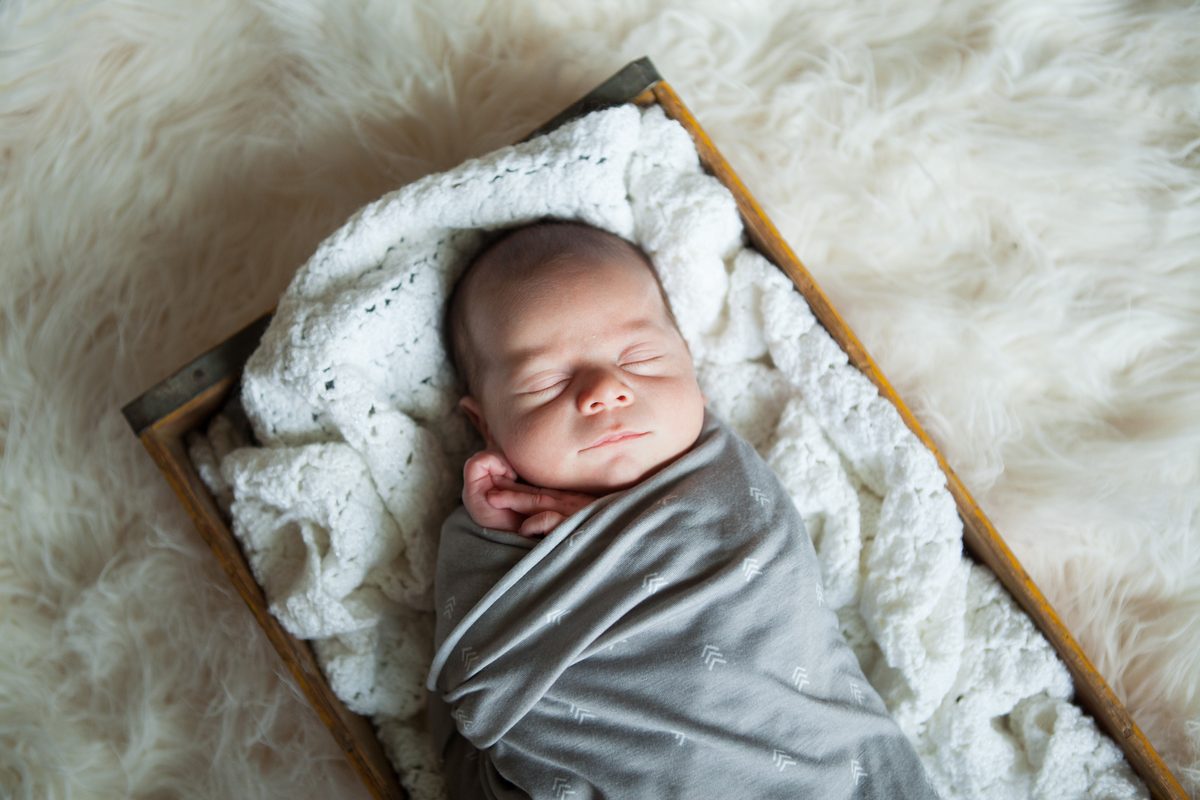A newborn in a gray swaddle sleeping in a crate of blankets